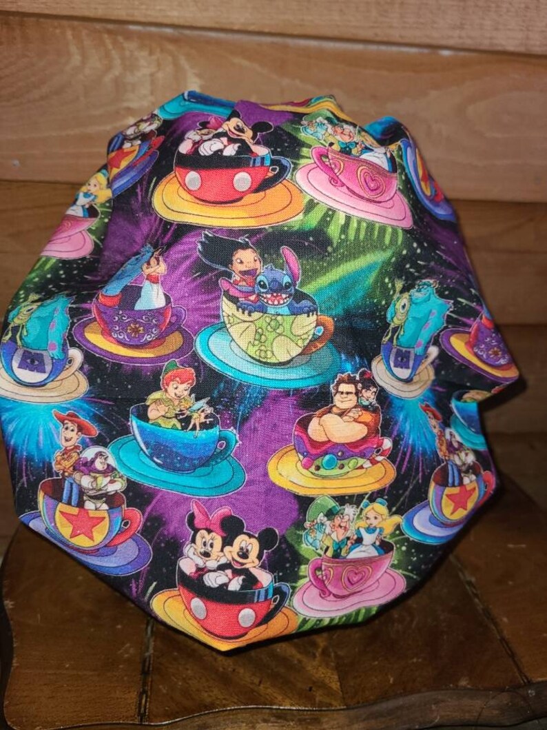 May include: A colorful fabric scrub hat with a black background and a galaxy print. The hat features various Disney characters riding in teacups, including Mickey Mouse, Minnie Mouse, Stitch, and Woody.