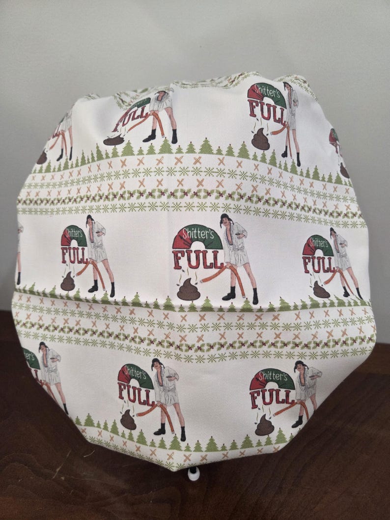May include: A white scrub cap featuring a repeating pattern of a person in a bathrobe holding a hose with the text Shitter's FULL above a brown pile. The design includes horizontal bands of green pine trees and small red berries on a white background.