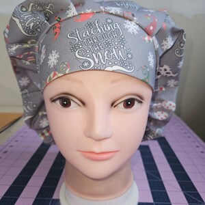 May include: Gray and white patterned surgical scrub hat with the text "Stacking Through The Snow" in white.