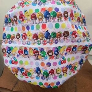 Pu&ograve; includere: Un berretto da lavoro in tessuto bianco con un motivo colorato di personaggi M&M's e pois. I personaggi sorridono e si tengono per mano. Lo sfondo &egrave; bianco con un motivo a coriandoli.
