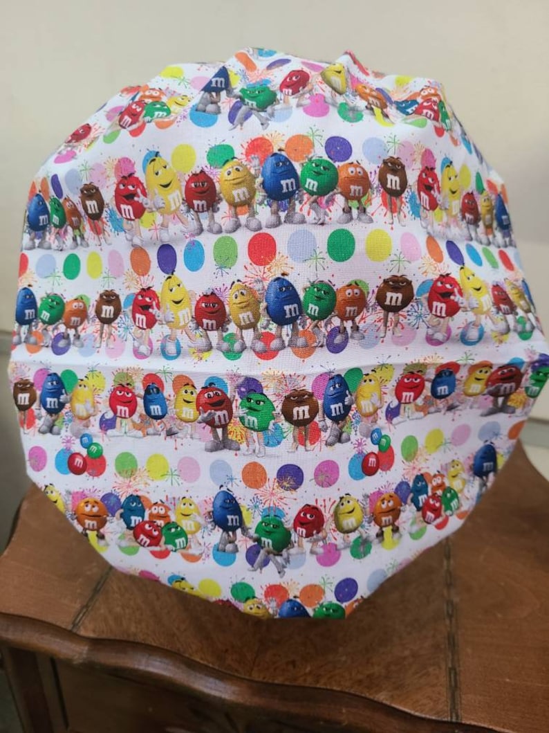 Pu&ograve; includere: Un berretto da lavoro in tessuto bianco con un motivo colorato di personaggi M&M's e pois. I personaggi sorridono e si tengono per mano. Lo sfondo &egrave; bianco con un motivo a coriandoli.