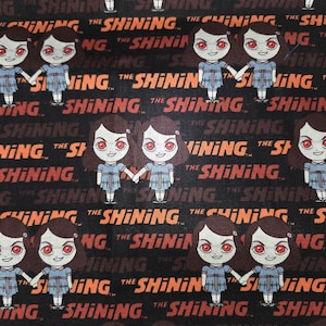May include: Black fabric featuring a repeating pattern of cartoon twins from "The Shining," holding hands. The text "THE SHINING" is printed in orange and red, creating a horror movie theme.