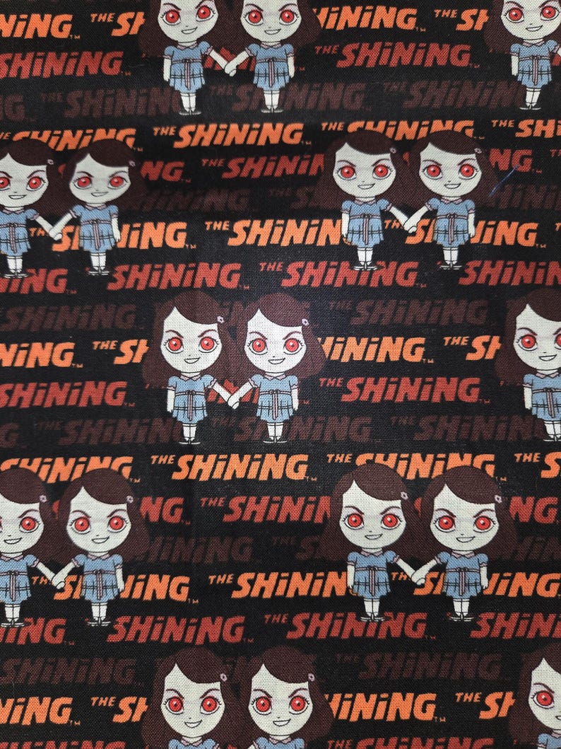 May include: Black fabric featuring a repeating pattern of cartoon twins from "The Shining," holding hands. The text "THE SHINING" is printed in orange and red, creating a horror movie theme.