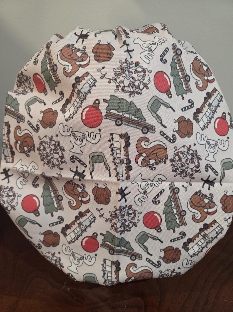 May include: A scrub cap featuring a cream-colored background with a repeating holiday pattern. The print includes brown squirrels wearing Santa hats, roasted turkeys, red ornaments, green Christmas trees on cars, campers, moose, and candy canes.