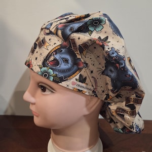 May include: A beige scrub cap featuring a repeating pattern of blue opossums, teal flowers, and gold banners with the text "AWESOME OPOSSUM". The fabric includes a small white tag with the brand name "ChristieStitches" in black script lettering.