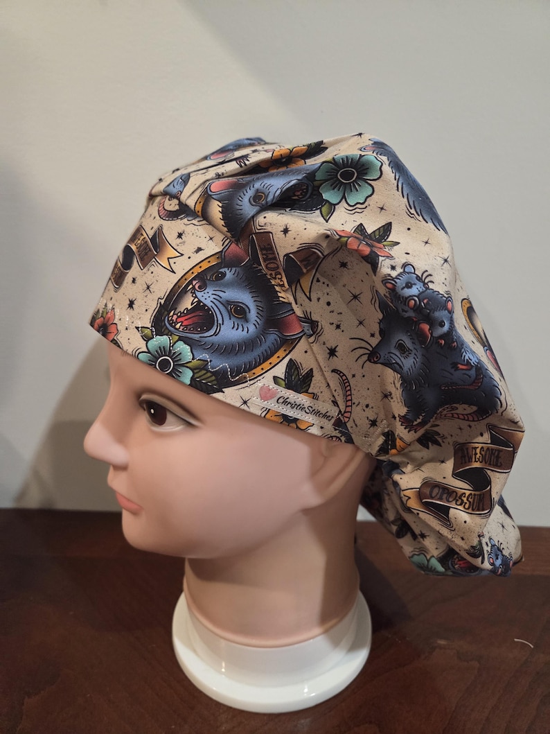 May include: A beige scrub cap featuring a repeating pattern of blue opossums, teal flowers, and gold banners with the text "AWESOME OPOSSUM". The fabric includes a small white tag with the brand name "ChristieStitches" in black script lettering.