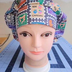 May include: A scrub hat with a colorful print of postage stamps featuring medical terms like "Cardiac", "General Surgery", and "Trauma".