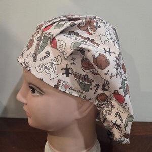 May include: A scrub cap with a white background featuring a repeating pattern of holiday-themed illustrations including moose, squirrels, candy canes, and vehicles. A small fabric tag with the text ChristieStitch is visible on the side.