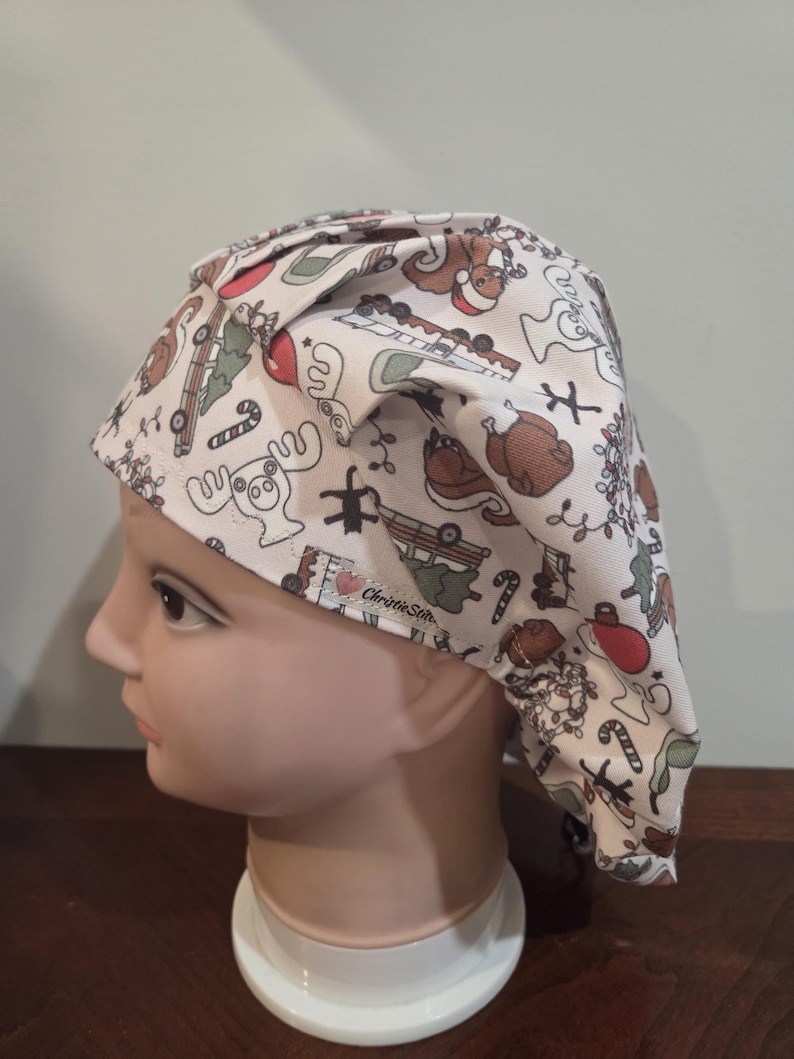 May include: A scrub cap with a white background featuring a repeating pattern of holiday-themed illustrations including moose, squirrels, candy canes, and vehicles. A small fabric tag with the text ChristieStitch is visible on the side.