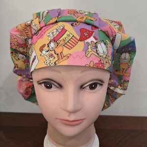 May include: A pleated scrub cap featuring a colorful cartoon print of Garfield the cat, popcorn buckets, and mushrooms on a background of pink, yellow, and green shapes. The fabric includes text that reads be a gentle person, be humble nor overbearing.