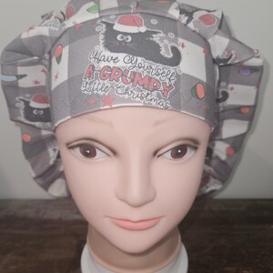 May include: A gray and white checkered scrub hat with a black cat wearing a Santa hat. The cat is surrounded by red and white stars and the text "Have yourself a grumpy little Christmas".