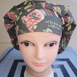 May include: A black and green patterned scrub hat with a watermelon and mason jar design. The hat has the text "cut the burn with a little lime" and "watermelon sunshine" printed on it.