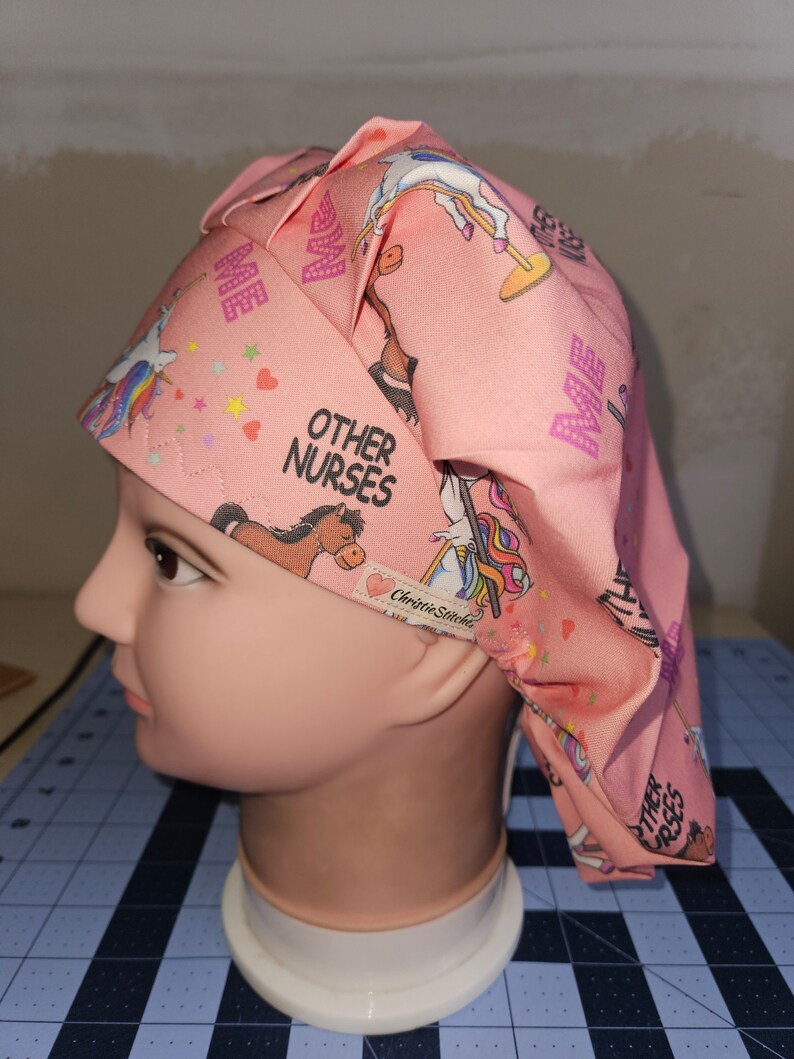 K&ouml;nnte beinhalten: Rosa chirurgische M&uuml;tze mit einem Einhorn- und Pferde-Druck. Die M&uuml;tze hat den Text "Other Nurses" und "Me" aufgedruckt. Die M&uuml;tze befindet sich auf einem wei&szlig;en Mannequin-Kopf.