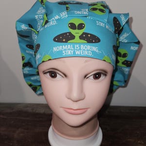 May include: A blue scrub cap with an alien print and the text "NORMAL IS BORING. STAY WEIRD." The cap features a repeating pattern of green aliens and the words "STAY WEIRD." The cap is displayed on a mannequin head.