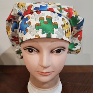 May include: A white surgical scrub cap featuring a colorful puzzle piece pattern in red, blue, green, and yellow. The cap is designed to cover the head and hair, commonly used in medical or culinary settings.