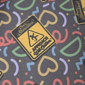 Può includere: Un tessuto grigio scuro con un motivo ripetuto di cartelli rettangolari gialli con il testo Over Stimulated e Approach With Caution, insieme a forme astratte colorate tra cui spirali rosse, semicerchi verdi e linee a zigzag azzurre.