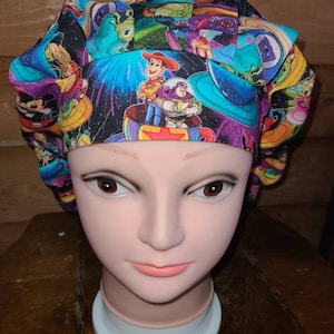 May include: A colorful surgical scrub hat with a space-themed print featuring characters from the Toy Story and Monsters Inc. movies. The print includes Woody, Buzz Lightyear, and Sulley.
