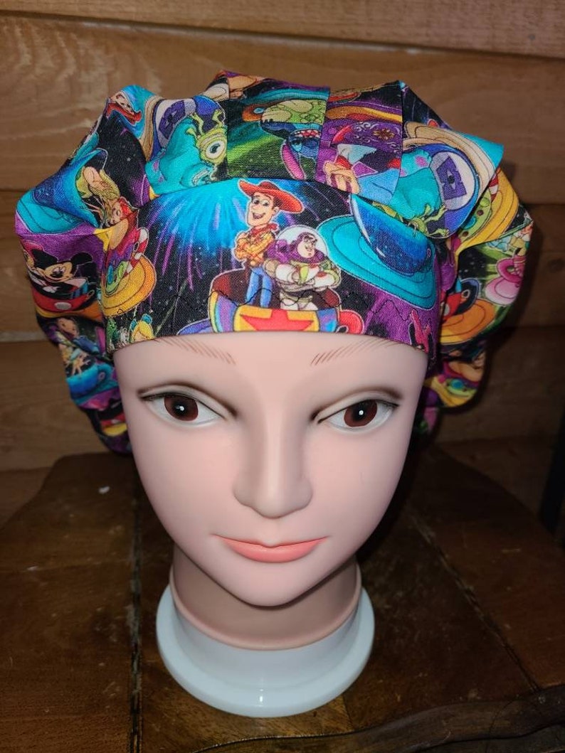 May include: A colorful surgical scrub hat with a space-themed print featuring characters from the Toy Story and Monsters Inc. movies. The print includes Woody, Buzz Lightyear, and Sulley.
