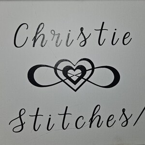 May include: A white canvas with black lettering that reads "Christie Stitches" and a black infinity symbol with a heart inside.