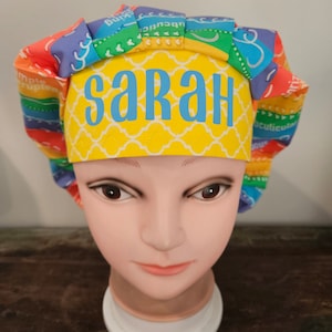 May include: A colorful scrub cap with the name "SARAH" in blue letters on a yellow background. The cap features a rainbow pattern and is displayed on a mannequin head.