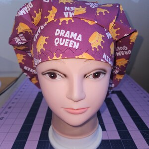 May include: A purple scrub cap with a repeating pattern of gold crowns and the words "DRAMA QUEEN" in white. The cap is displayed on a mannequin head.