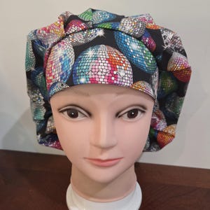 May include: A colorful bouffant surgical scrub cap featuring a repeating pattern of disco balls in shades of blue, green, pink, and orange on a black background. The cap is displayed on a mannequin head.