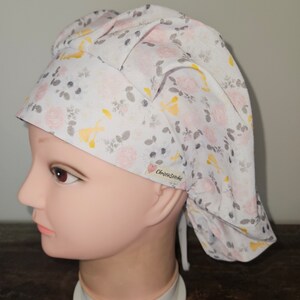 May include: A white surgical scrub cap with a floral pattern of pink roses, yellow accents, and gray leaves. The cap is tied at the back and features the text "ChristieStitches" on a small tag.