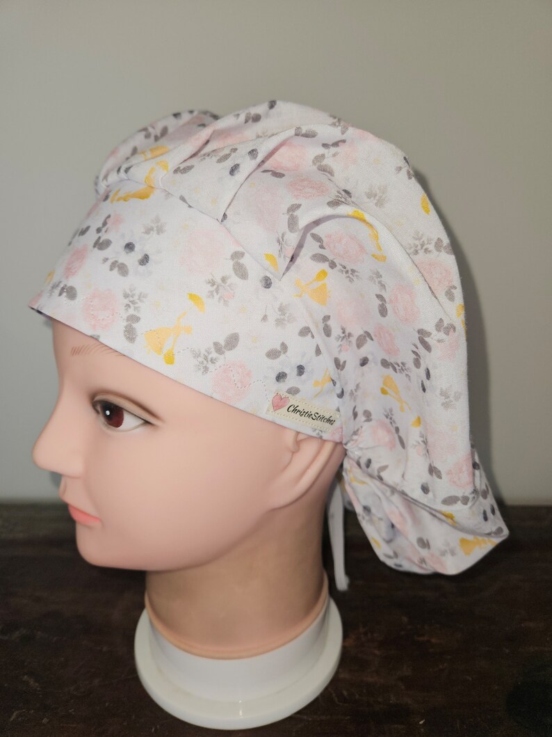 May include: A white surgical scrub cap with a floral pattern of pink roses, yellow accents, and gray leaves. The cap is tied at the back and features the text "ChristieStitches" on a small tag.
