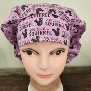 May include: A lavender scrub cap featuring a repeating pattern of black squirrel silhouettes and the text "Oh look a SQUIRREL" printed in black. The cap has a pleated front band and a gathered back section, constructed from fabric with a visible zigzag stitch.