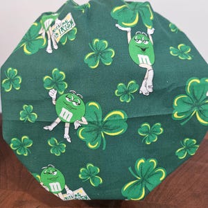 Pu&ograve; includere: Un berretto da scrub in tessuto verde con un motivo ripetuto di trifogli verdi e personaggi M&M verdi. I personaggi M&M indossano scarpe bianche e tengono trifogli. Un personaggio M&M tiene un cartello che dice "Kiss Me I'm Irish".