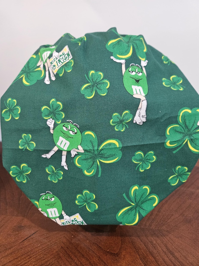 Pu&ograve; includere: Un berretto da scrub in tessuto verde con un motivo ripetuto di trifogli verdi e personaggi M&M verdi. I personaggi M&M indossano scarpe bianche e tengono trifogli. Un personaggio M&M tiene un cartello che dice "Kiss Me I'm Irish".