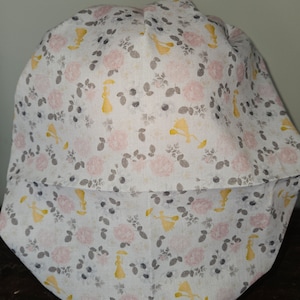 May include: A white fabric hat with a floral pattern featuring pink roses, gray leaves, and yellow figures. The hat has a brim and a rounded crown. The overall design is soft and feminine.