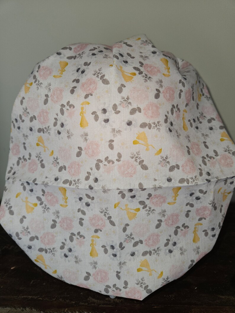 May include: A white fabric hat with a floral pattern featuring pink roses, gray leaves, and yellow figures. The hat has a brim and a rounded crown. The overall design is soft and feminine.
