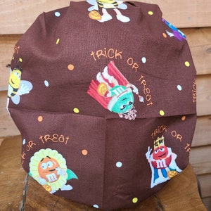 Pu&ograve; includere: Un cappello da lavoro in tessuto marrone a tema Halloween. Il cappello presenta un motivo ripetitivo di personaggi M&M's vestiti in costume, tra cui un'ape, un re, un girasole e un personaggio di mais dolce. Il testo "trick or treat" &egrave; ripetuto in tutto il motivo.