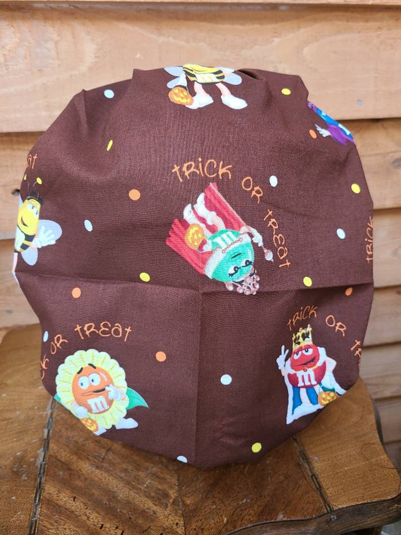 Pu&ograve; includere: Un cappello da lavoro in tessuto marrone a tema Halloween. Il cappello presenta un motivo ripetitivo di personaggi M&M's vestiti in costume, tra cui un'ape, un re, un girasole e un personaggio di mais dolce. Il testo "trick or treat" &egrave; ripetuto in tutto il motivo.