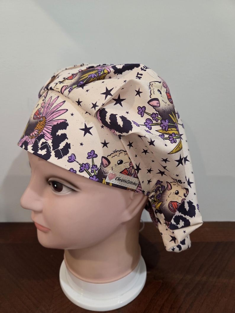 May include: A cream-colored scrub cap featuring a repeating pattern of opossums, purple lavender sprigs, black stars, and leopard print accents. The fabric cap includes a small white tag with the text ChristieStitches and a pink heart icon.