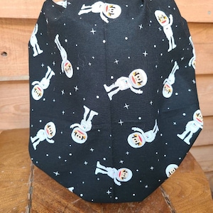 May include: Black scrub hat with a repeating pattern of white astronauts on a black background with white stars.