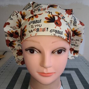 May include: A white scrub hat with a turkey print and the text "Gobble Til You Wobble!" in brown lettering. The hat is on a mannequin head.