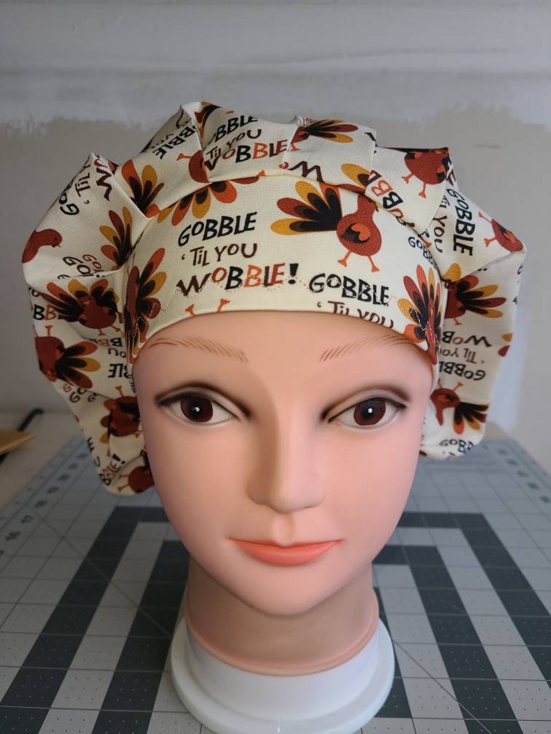 May include: A white scrub hat with a turkey print and the text "Gobble Til You Wobble!" in brown lettering. The hat is on a mannequin head.