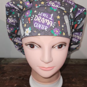 May include: A gray scrub hat with a purple and white floral print. The hat has the text "Level 1 Drama Center" in a purple circle.