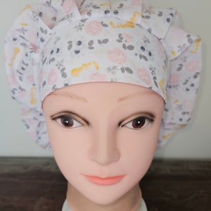 May include: A white bouffant surgical scrub cap with a floral pattern of pink roses, yellow accents, and gray leaves. The cap is gathered at the back and has a comfortable fit. The cap is designed for medical professionals.