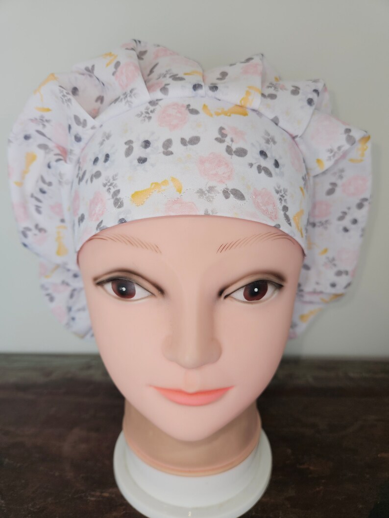 May include: A white bouffant surgical scrub cap with a floral pattern of pink roses, yellow accents, and gray leaves. The cap is gathered at the back and has a comfortable fit. The cap is designed for medical professionals.