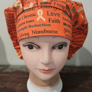 May include: An orange scrub cap featuring a repeating pattern of awareness ribbons and printed words including Chronic, Love, Faith, Strength, Reduction, Numbness, Suffering, Spasms, and Balance. The fabric has a pleated front band.