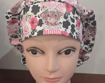 May include: A pleated scrub cap featuring a white background with black cow spots, pink floral patterns, and a central illustration of a Highland cow wearing a flower crown. The text "Not in the MOOD" is printed in pink above and below the cow.