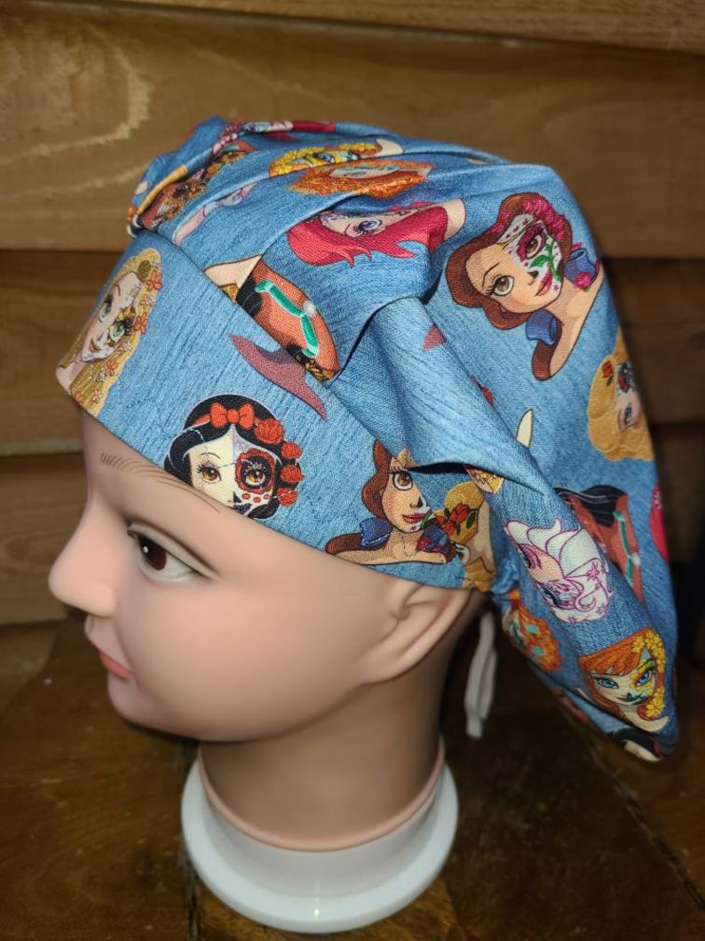 May include: A blue scrub hat with a print of various Disney princesses with Day of the Dead makeup. The hat has a white drawstring.