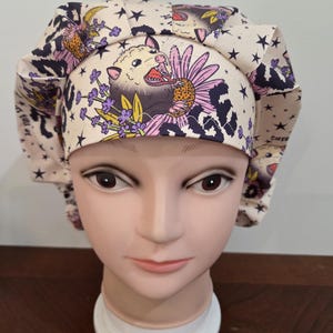 May include: A scrub cap in a cream color featuring a repeating pattern of opossums, purple lavender sprigs, pink flowers, and black stars. The fabric has a pleated design around the crown and includes partial text visible on the side.