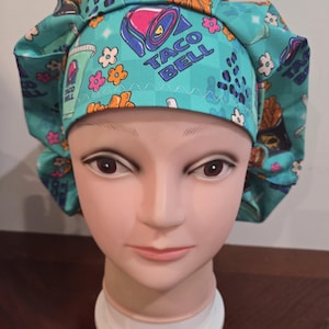 May include: A teal scrub cap featuring a repeating pattern of Taco Bell logos, drink cups, and colorful flowers. The fabric has a subtle checkered background with blue leopard print accents. The cap is gathered with a band across the forehead.