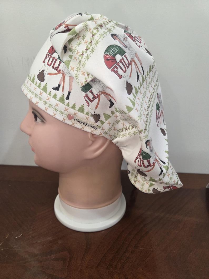 May include: A white scrub cap featuring a repeating Christmas-themed pattern with green pine trees, brown poop emojis, and text that reads "Shitter's Full". The fabric also includes the brand name "ChristieStitcher" with a small red heart icon.