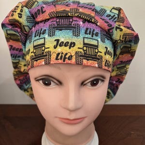 May include: A colorful bouffant scrub hat with a rainbow gradient background. The hat features black Jeep vehicles and the words "Jeep Life" in a repeating pattern. The hat is displayed on a mannequin head.