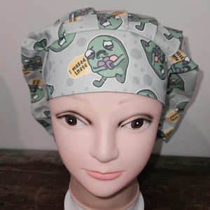 May include: A light green scrub hat with a repeating pattern of green cartoon characters holding purple hearts. The characters have a speech bubble that says "I maked these".